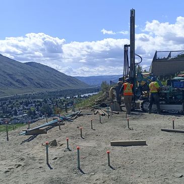 Construction workers operate machinery on a hillside overlooking a valley with houses and a river.