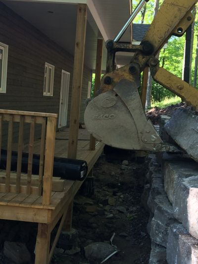 Excavator bucket aligning an armour stone onto top of retaining wall directly beside a porch.