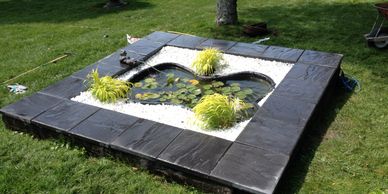 Small kidney shaped backyard pond surrounded in small white stone, plants and large dark stone 