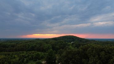 Sunset peeks through dark clouds over a lush, forested hill and rural homes.