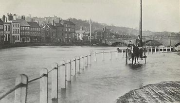 Historic black-and-white photo of a horse-drawn cart on a flooded street by riverside buildings.