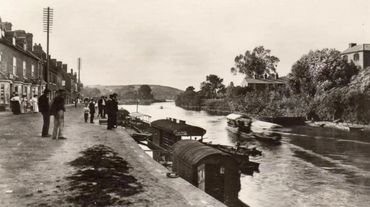 Vintage riverside scene with people, boats, and old buildings along the shore.