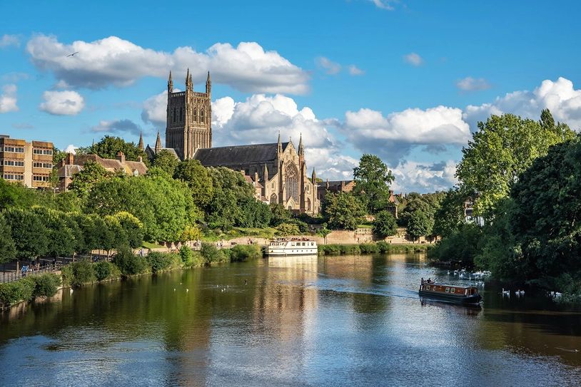 Scenic riverside view with historic cathedral, boats, and lush greenery under a blue sky.