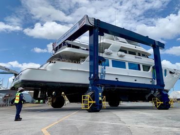 A large yacht lifted by a blue marina crane with a worker nearby.