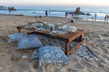 bohemian beach picnic dining setup with pillow seats and a wooden table at sunset on the sand