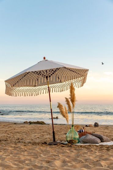 A bohemian style umbrella stands over a picnic blanket, basket and pillows with vases at sunset