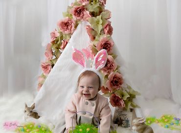 6 month baby girl posing by the tee pee draped in pink flowers , she's wearing a bunny cap