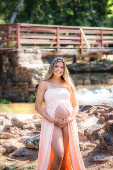 Pregnant woman in a flowing peach dress smiles by a rocky riverside with a wooden bridge behind.