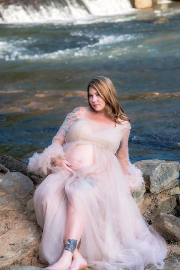Pregnant woman in a flowing dress poses by a river on rocks.