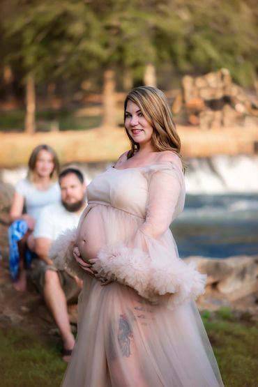 Pregnant woman in a sheer gown smiling outdoors with couple in background.