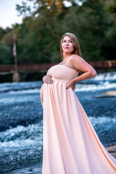 Pregnant woman in a flowing peach dress stands confidently by a river.