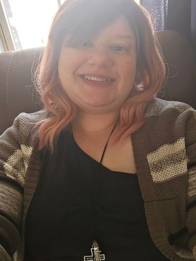 Smiling woman with pink hair wearing a striped cardigan and cross necklace.