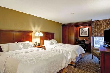 Hotel room with two queen beds, wooden furniture, and a window with patterned curtains.