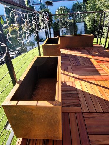 Sunlit balcony with empty planters and wooden flooring overlooking a garden and houses.