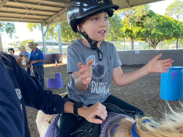 A young boy wearing a helmet rides a horse with support during a therapeutic riding session.