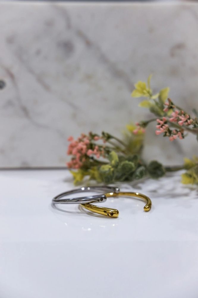 Two minimalist metal rings, one silver and one gold, on a white surface with blurred flowers in the background.