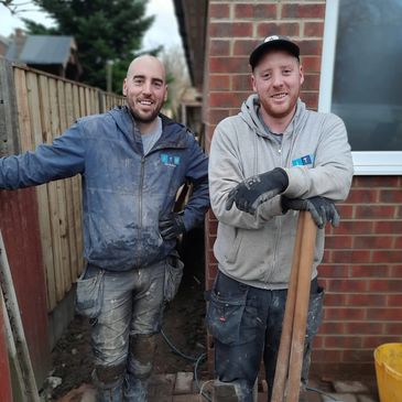 Two muddy construction workers posing with tools outside a brick building.