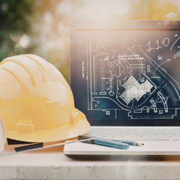 Yellow construction helmet and blueprints beside a laptop displaying architectural plans.