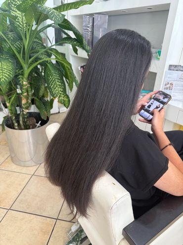 Person with long, straight black hair using a phone, next to a potted plant.