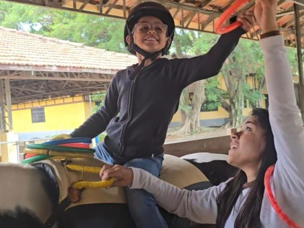 Criança sorridente montando em um touro mecânico, com ajuda de uma mulher.