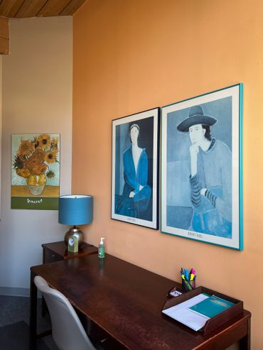 A cozy office corner with art prints, a wooden desk, and blue lamp.