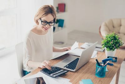 Woman using calculator and laptop at a desk in a bright office.