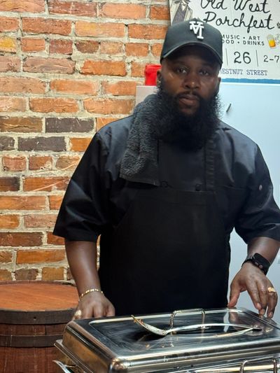 A chef with a neatly groomed beard stands behind a food tray in a rustic setting, surrounded by warm wooden textures and natural lighting. He appears focused and composed, presenting the dish with confidence and professionalism. The scene highlights a blend of culinary skill and down‑to‑earth charm.