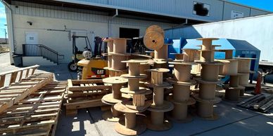 Wooden spools stacked outdoors near a warehouse and forklift.