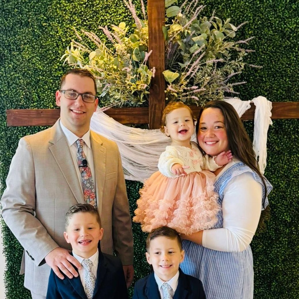 A happy family of five posing in front of a decorative cross with greenery.
