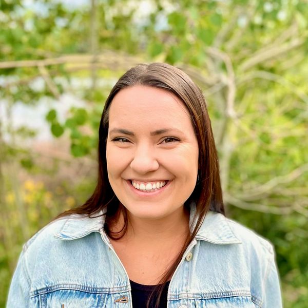 A smiling woman in a denim jacket outdoors with greenery in the background.