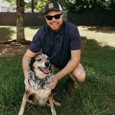 Man with sunglasses and cap smiling while holding a happy dog outdoors.
