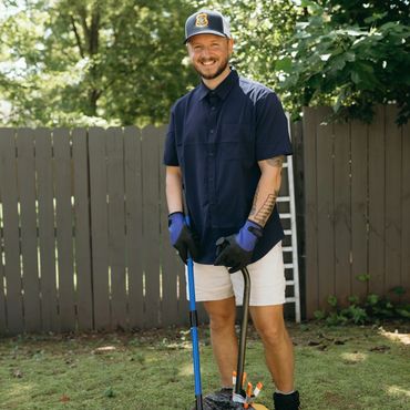 Smiling man in gardening gloves holding a broom and dustpan outdoors.