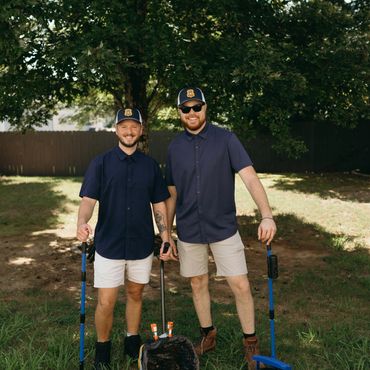 Two men in navy shirts and caps holding cleaning tools outdoors.