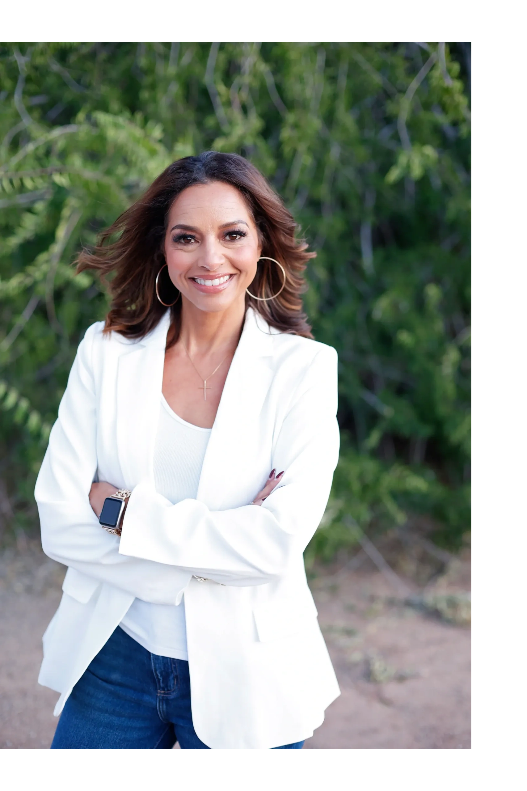 Confident woman in white blazer smiling outdoors with arms crossed.
