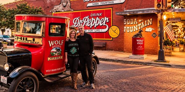 Couple posing by vintage Wolf Brand Chili truck near Dr Pepper soda fountain.