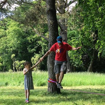a young lady helping her dad on a slackline in the woods