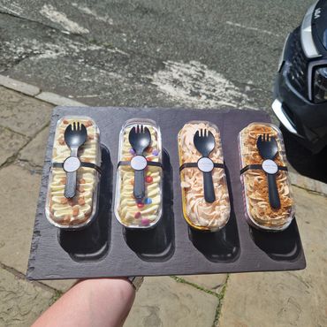Four decorated dessert containers with spoons on a black tray outdoors.