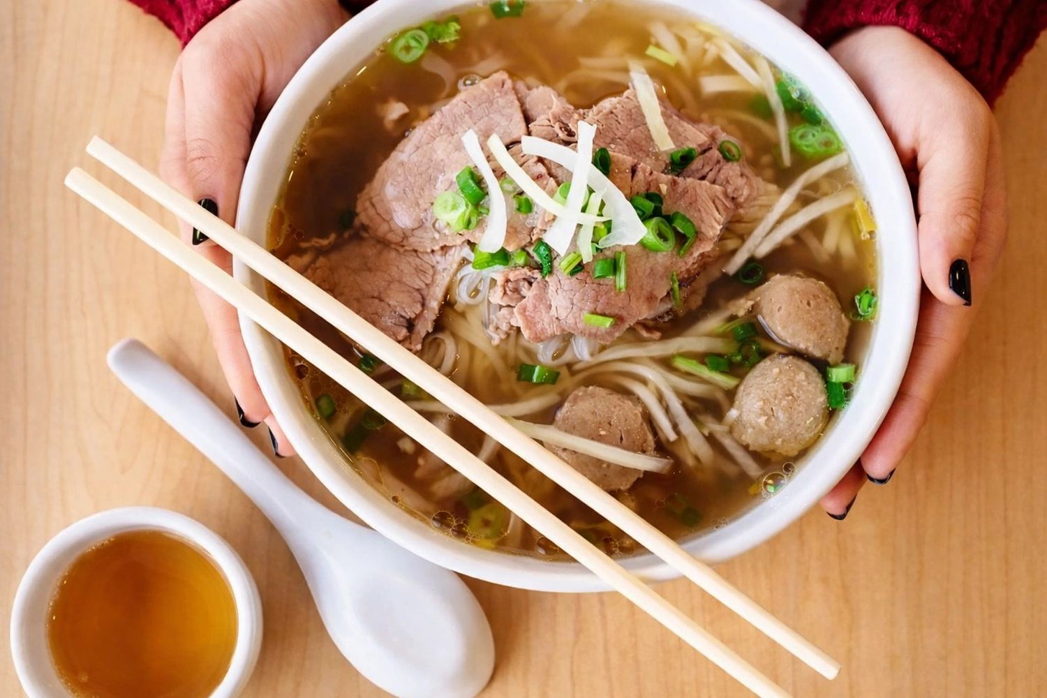 Hands holding a bowl of pho with chopsticks and tea.