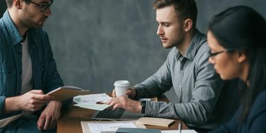 Three professionals engaged in a focused meeting around a wooden table with documents and laptops.