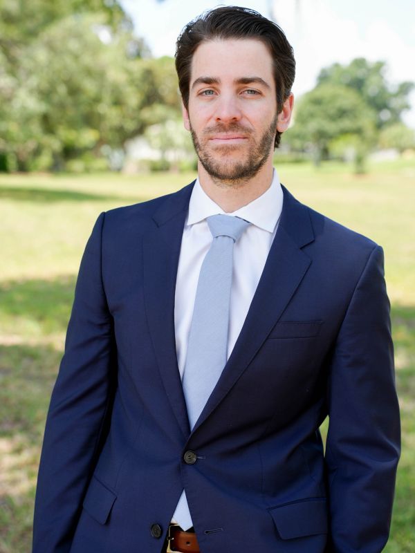 Man in a navy blue suit and light blue tie standing outdoors.