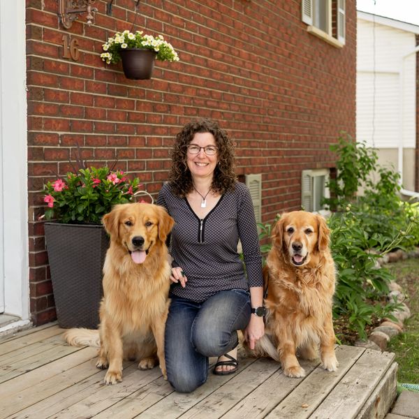 A smiling woman kneels with two golden retrievers on a wooden porch outside a brick house.