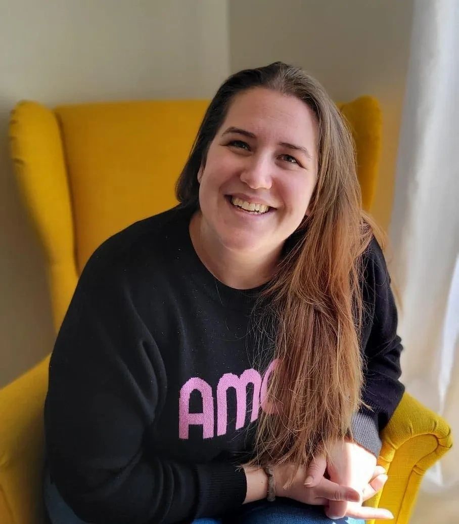 Smiling woman with long hair sitting on a yellow chair in cozy indoor lighting.