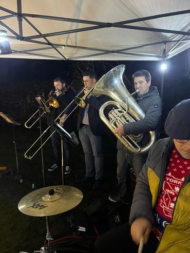 A brass band performs under a tent at night with trombones and a tuba.