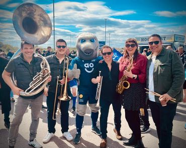 Group of musicians posing with a sports mascot outdoors on a sunny day.