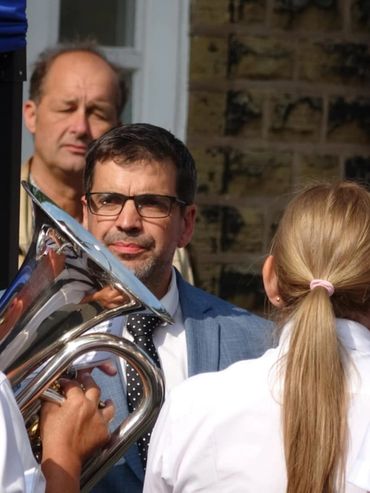A group of musicians playing brass instruments outdoors, with a focused man in a blue suit.