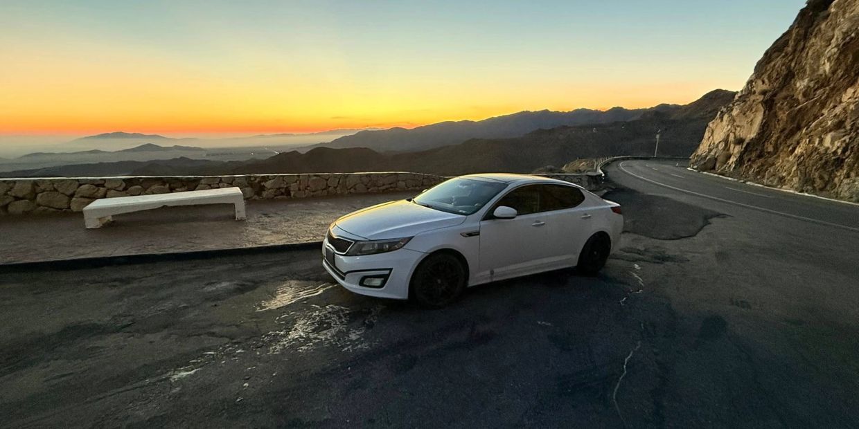 White sedan parked near a mountain road at sunset with a stone bench nearby.