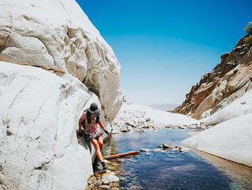 A person hiking near a stream between large white rocks under a clear blue sky.