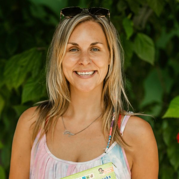 Smiling woman with blonde hair holding colorful books against green foliage.