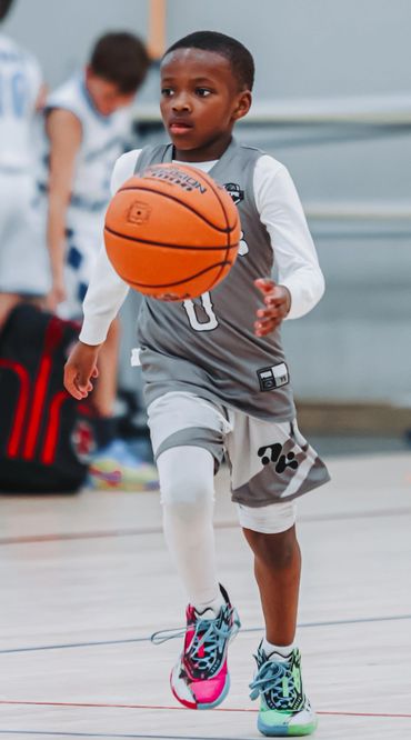 Young boy in gray basketball uniform dribbling a basketball indoors.