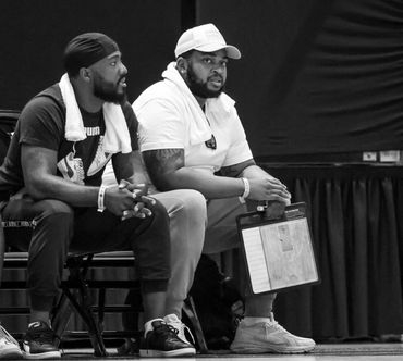 Two men sitting with towels around their necks, one holding a basketball strategy board.